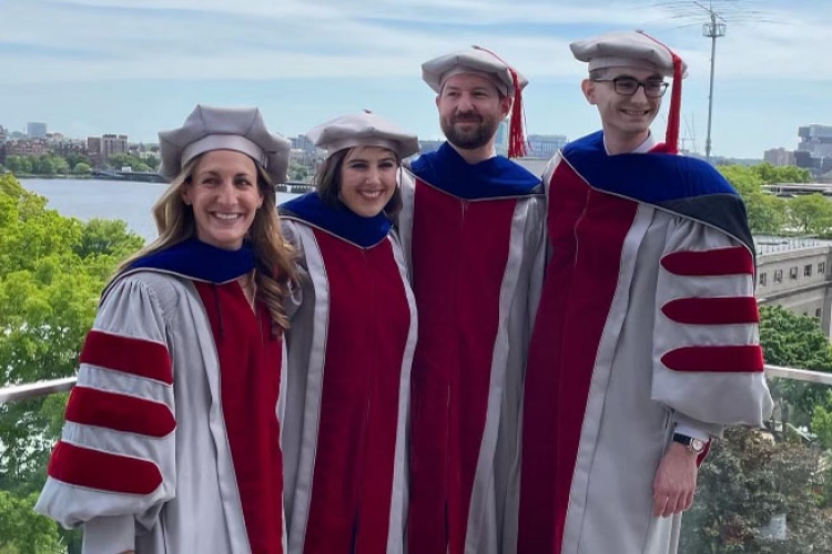 Four graduates of MIT SSP's PhD program stand on a balcony in full graduation regalia