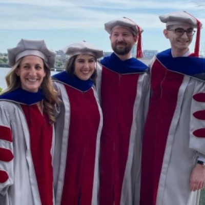 Four graduates of MIT SSP&#039;s PhD program stand on a balcony in full graduation regalia